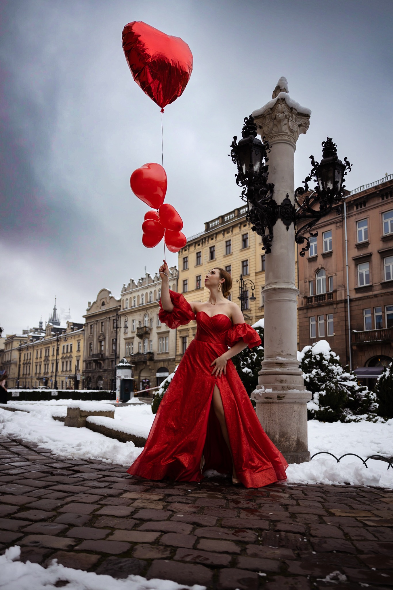 Kobieta w czerwonej, błyszczącej sukni pozuje z czerwonymi balonami w kształcie serca na tle zabytkowej architektury i ośnieżonego bruku.