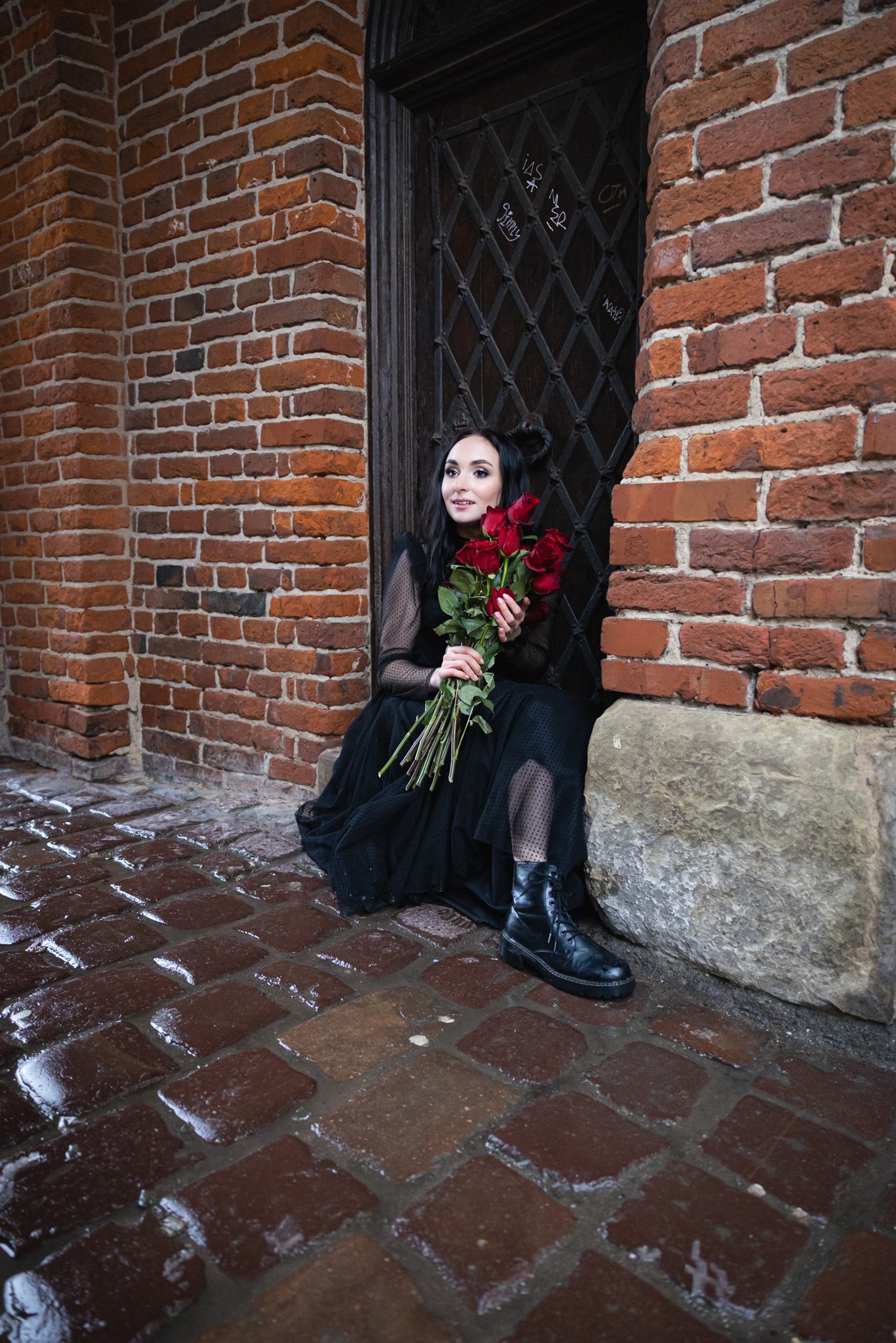 Kobieta w czarnej sukience i butach siedzi na mokrym bruku przy ceglanej ścianie i drzwiach z kratą, trzymając bukiet czerwonych róż.