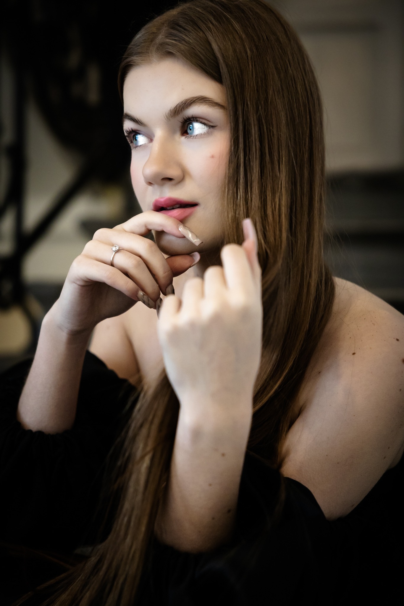 Portret młodej kobiety z długimi brązowymi włosami, zamyślona, z pierścionkiem zaręczynowym na palcu, w czarnej bluzce z odkrytymi ramionami, spoglądająca w bok.