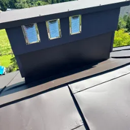 Nowoczesny komin wentylacyjny w kolorze grafitowym z trzema metalowymi kratkami wentylacyjnymi, zamontowany na brązowym dachu z blachy na tle zieleni drzew i wzgórz.