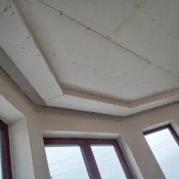 Sufit z zamontowaną płytą kartonowo-gipsową, widoczne śruby mocujące, surowe wykończenie, okna z brązowymi ramami w tle.