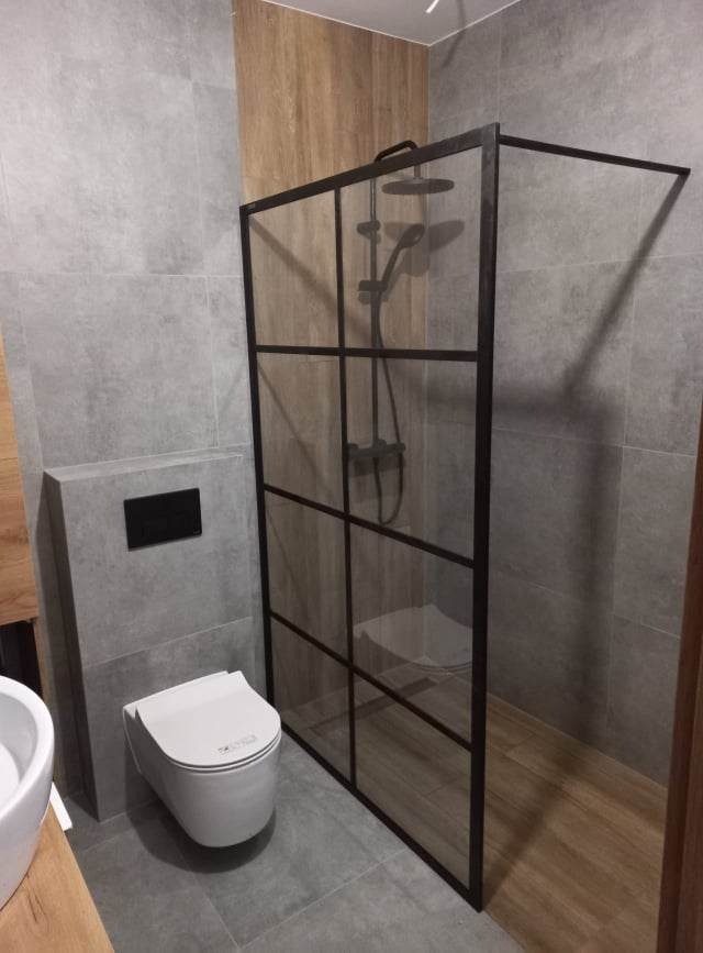 Nowoczesna łazienka z kabiną prysznicową typu walk-in z czarnymi profilami, wiszącą miską WC i szarą płytką imitującą kamień na ścianach i podłodze.