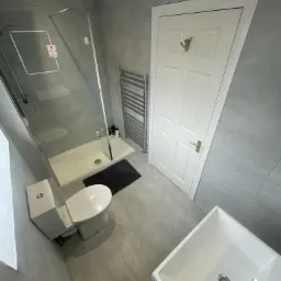 Nowoczesna łazienka z szarymi płytkami, kabiną prysznicową ze szklanymi drzwiami z naklejką informacyjną, białą toaletą, grzejnikiem drabinkowym i umywalką wpuszczaną w blat.