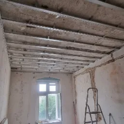 Montaż metalowej konstrukcji sufitu podwieszanego w pokoju z oknem i dwiema drabinami, ściany w trakcie remontu, widoczne ślady po instalacjach elektrycznych.