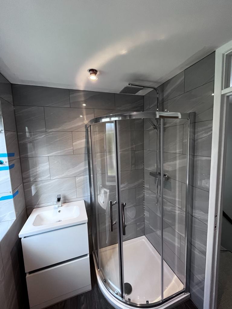 Nowoczesna łazienka po remoncie: kabina prysznicowa półokrągła z chromowanymi detalami, biała szafka z umywalką, szare płytki imitujące marmur na ścianach, ciemna podłoga, oświetlenie punktowe.