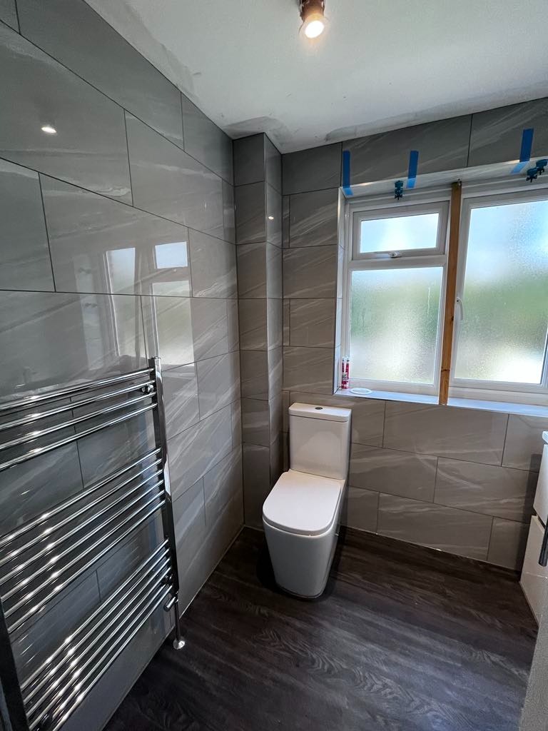 Wyremontowana łazienka w szarościach z ciemną podłogą, widoczna toaleta, grzejnik drabinkowy i okno z niebieską taśmą malarską na ramie.