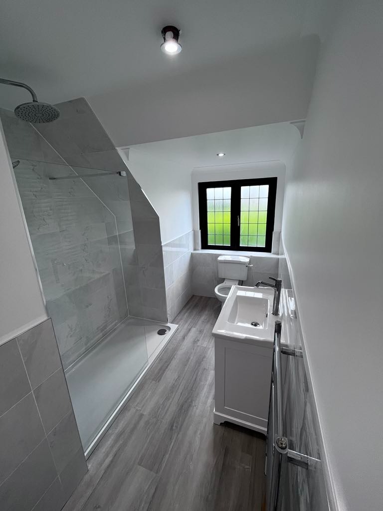 Nowoczesna łazienka z kabiną prysznicową typu walk-in, umywalką w białej szafce i grzejnikiem drabinkowym, wykończona szarymi płytkami imitującymi drewno na podłodze i szarymi płytkami na ścianach,...