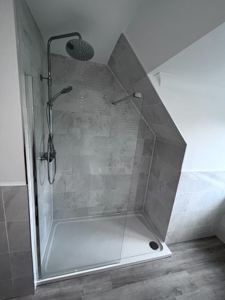 Nowoczesna kabina prysznicowa z deszczownicą i szklaną ścianką, wykończona szarymi płytkami imitującymi marmur, na podłodze panele w odcieniach szarości.