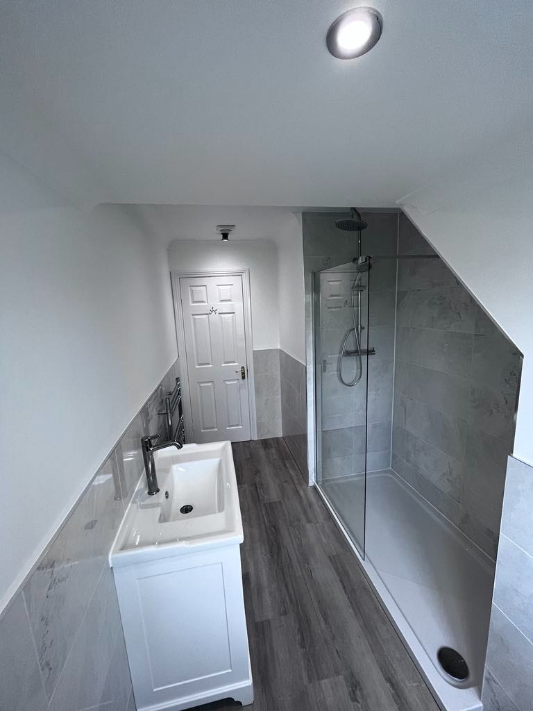 Nowoczesna łazienka w odcieniach szarości i bieli, z kabiną prysznicową ze szklanymi drzwiami, umywalką na szafce i drewnopodobną podłogą, oświetlona punktowo.