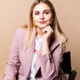 Portret kobiety z blond włosami w różowym żakiecie i koszulce z nadrukiem, pozującej na beżowym tle z dłonią pod brodą i złotymi kolczykami.