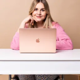 Uśmiechnięta blondynka w różowej bluzie siedzi przy białym biurku z laptopem w kolorze różowego złota, opierając brodę na dłoni z pomalowanymi na różne kolory paznokciami.