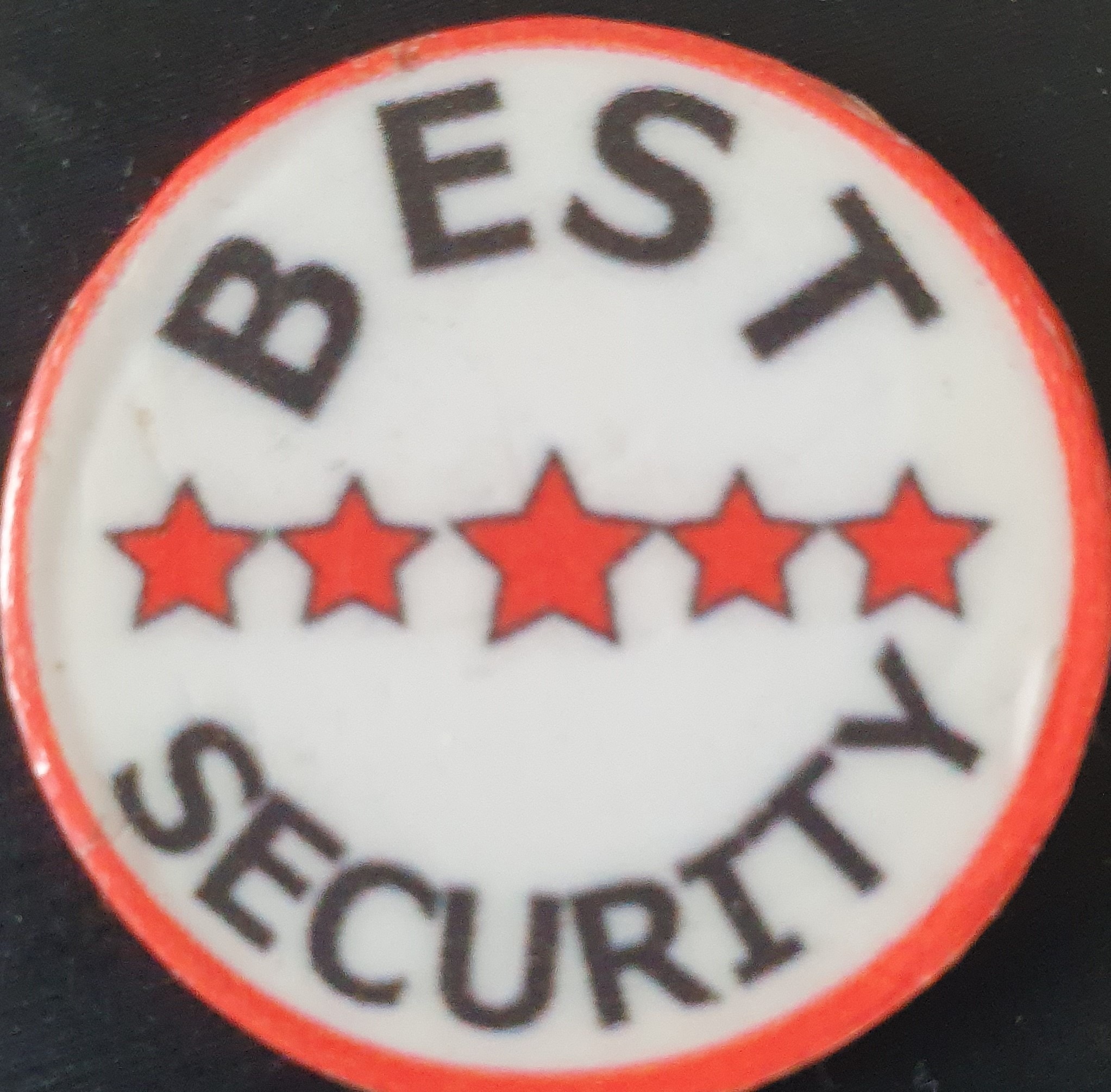 Okrągła odznaka z napisem 'BEST SECURITY' i pięcioma czerwonymi gwiazdkami na białym tle z pomarańczową obwódką.