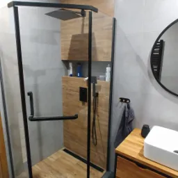 Nowoczesna łazienka z kabiną prysznicową typu walk-in z czarnymi profilami, drewnianą zabudową z półkami wewnątrz prysznica, umywalką nablatową na drewnianej szafce i okrągłym lustrem.