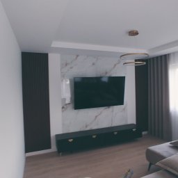 Rem Kompleks - Nowoczesny salon z telewizorem na ścianie z marmurowym wzorem, pod nim czarna szafka RTV z złotymi uchwytami, designerska lampa wisząca i szare zasłony.