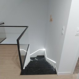 Rem Kompleks - Nowoczesne schody z czarnego marmuru z podświetleniem LED, szklana balustrada z czarną ramą, widok z góry na jasną podłogę.