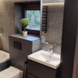 Nowoczesna łazienka z szarymi płytkami, podwieszaną toaletą, umywalką z szafką w kolorze antracytowym i dekoracyjnym drzewkiem w doniczce na zabudowie stelaża.