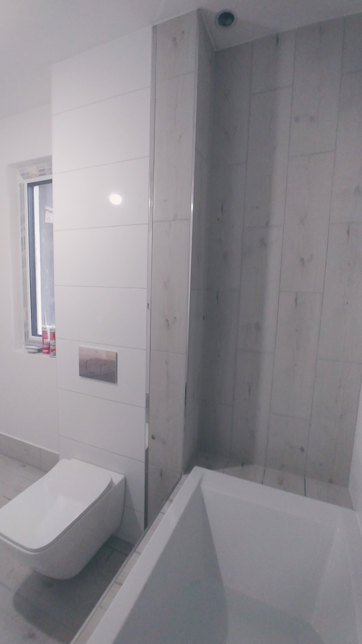 Nowoczesna łazienka w odcieniach bieli i szarości z wiszącą toaletą, zabudową z płytek i wanną, widoczne silikony przy oknie.