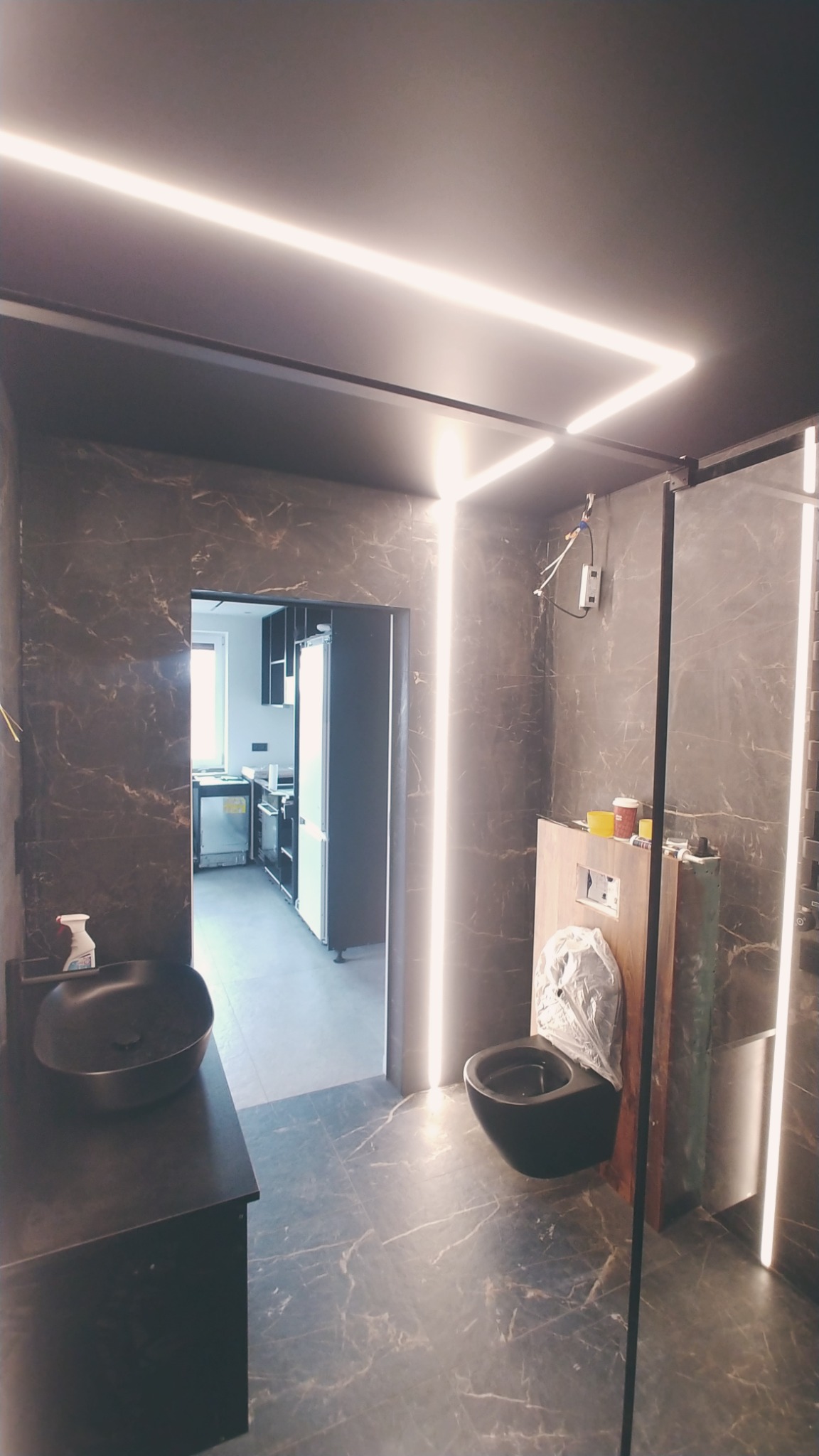 Nowoczesna łazienka z czarnymi marmurowymi płytkami, podwieszaną toaletą w folii ochronnej i liniowym oświetleniem LED na suficie, widok na kuchnię w tle.