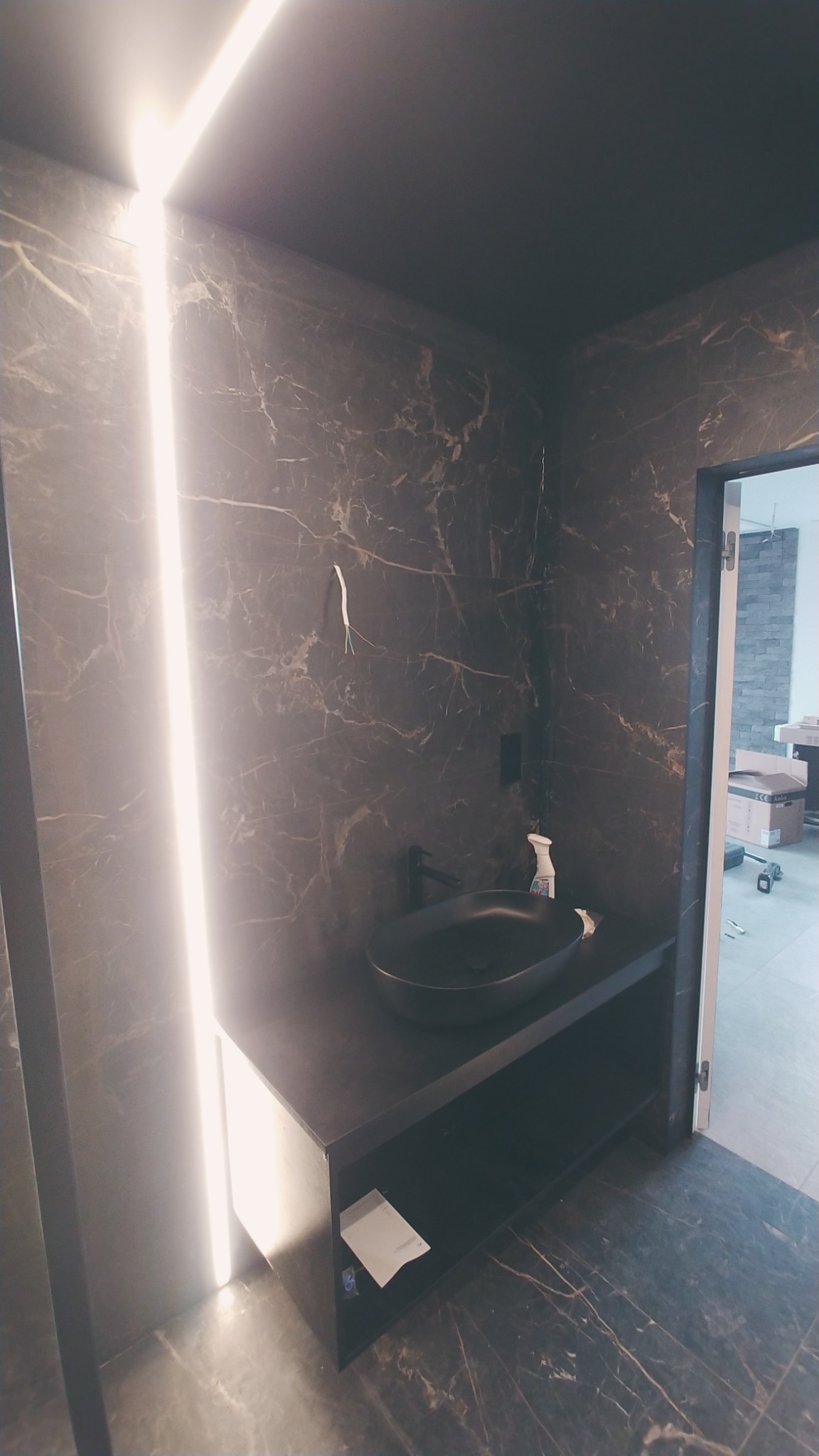 Nowoczesna łazienka z czarnymi marmurowymi płytkami, podświetlanym pionowym paskiem LED, czarną umywalką nablatową i otwartą szafką pod umywalką z widocznym fragmentem instalacji elektrycznej.