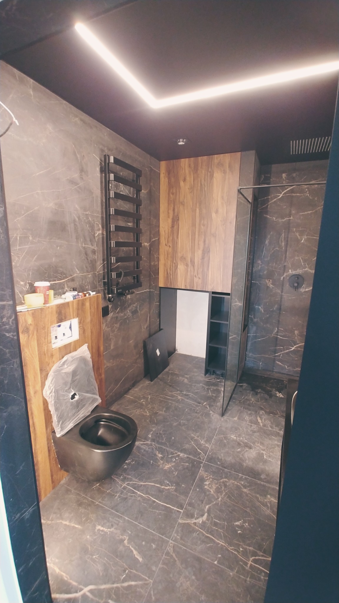 Nowoczesna łazienka z ciemnymi marmurowymi płytkami, podwieszaną toaletą z drewnianą obudową i grzejnikiem drabinkowym, oświetlona liniowym światłem LED na suficie.