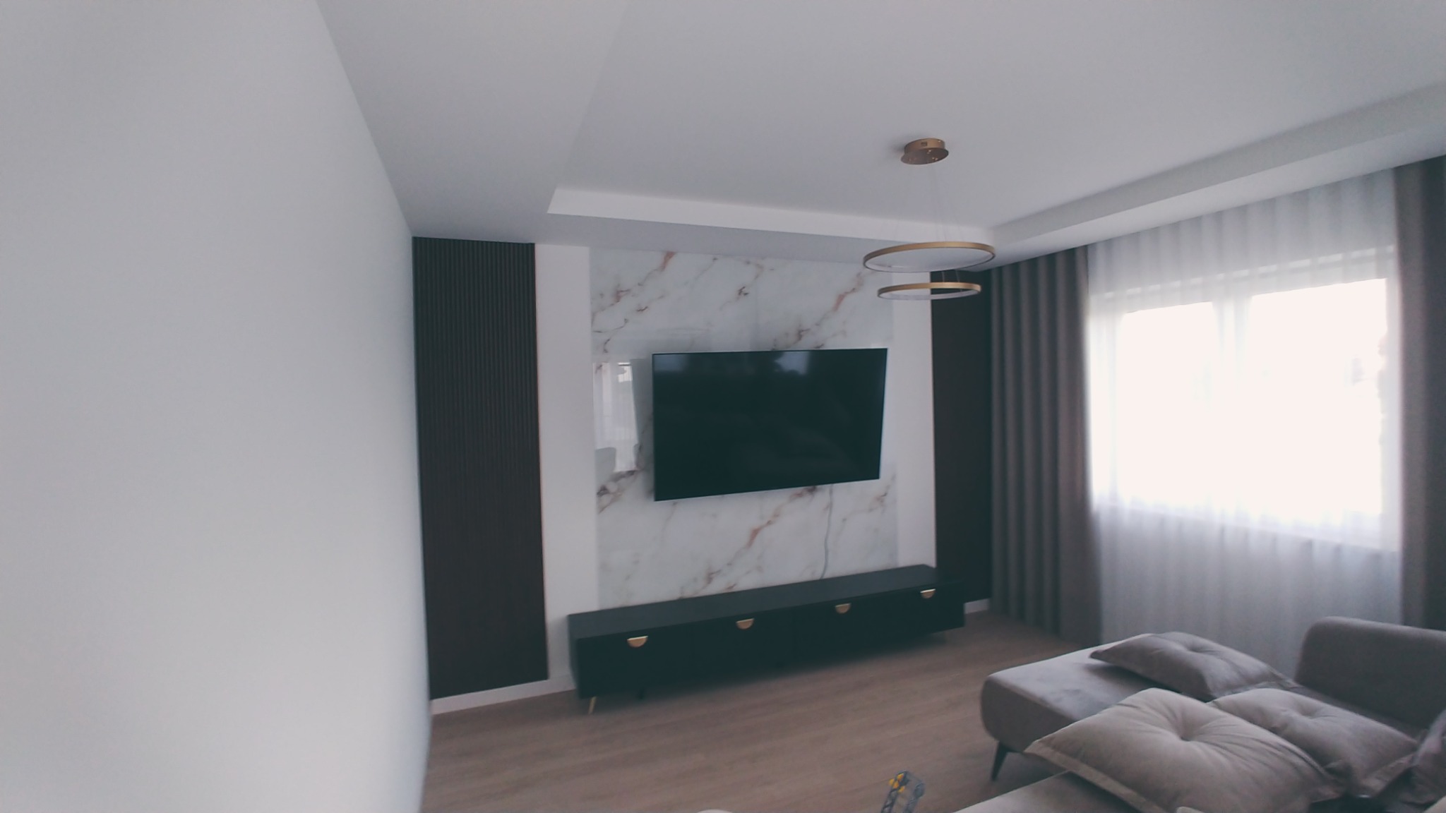 Nowoczesny salon z telewizorem na ścianie z marmurowym wzorem, pod nim czarna szafka RTV z złotymi uchwytami, designerska lampa wisząca i szare zasłony.