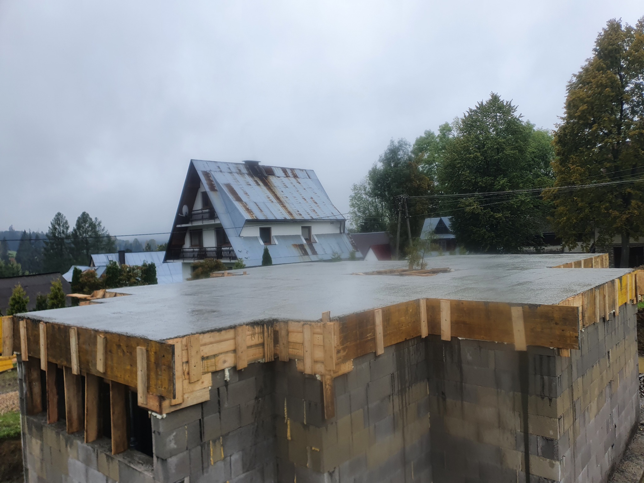 Wylany beton na stropie domu w trakcie budowy, widoczne szalunki drewniane, bloczki betonowe i otoczenie z innymi budynkami w tle, pochmurny dzień.