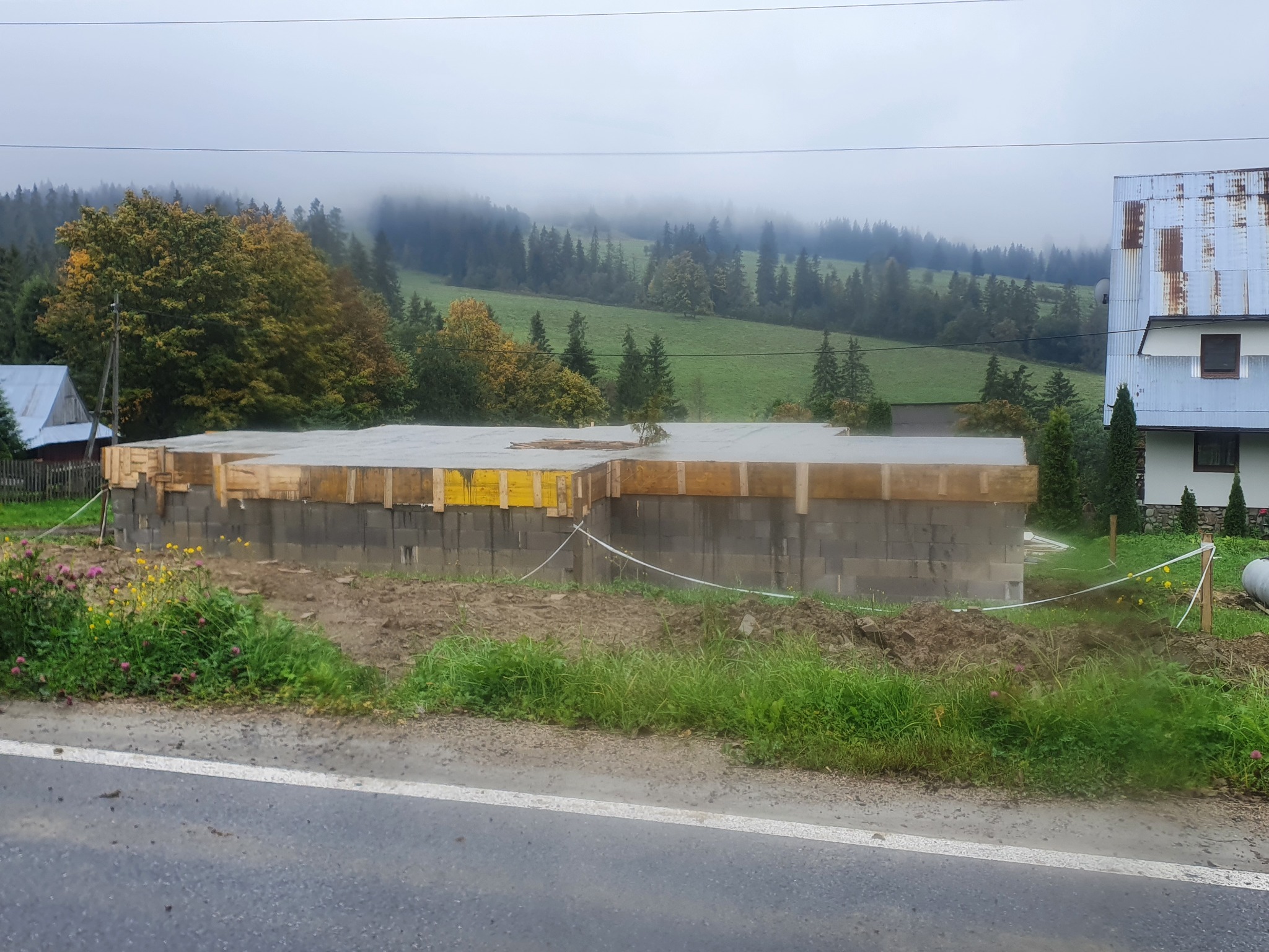 Wylany fundament z betonowych bloczków i drewnianym szalunkiem na tle mglistego, zalesionego wzgórza i częściowo widocznego domu z blachy falistej.