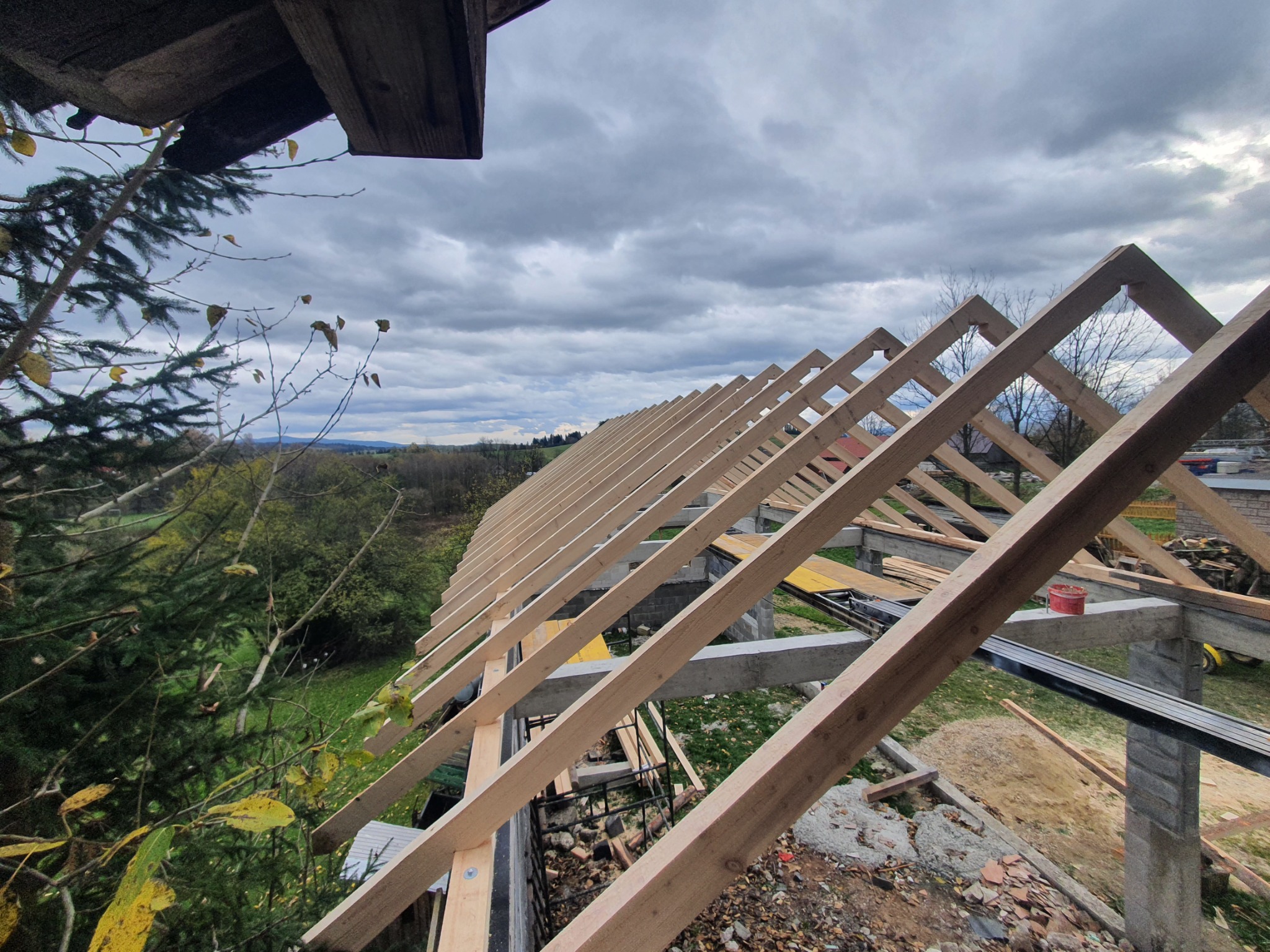 Konstrukcja więźby dachowej z drewnianych krokwi, widok z góry na surowy szkielet dachu, z betonowymi filarami i widokiem na zielone drzewa i pochmurne niebo w tle.