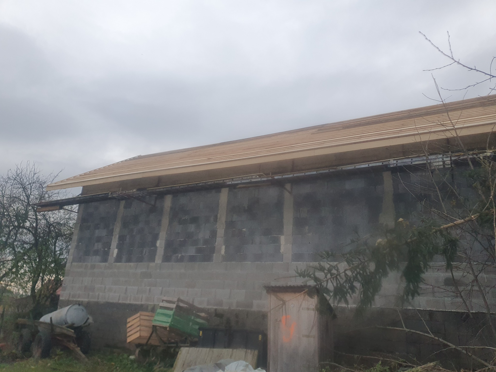 Surowy budynek z szarego betonu komórkowego z widocznym szkieletem dachu z drewnianych łat, przy pochmurnym niebie. Na pierwszym planie widoczne pozostałości po budowie, w tym beczka i palety.