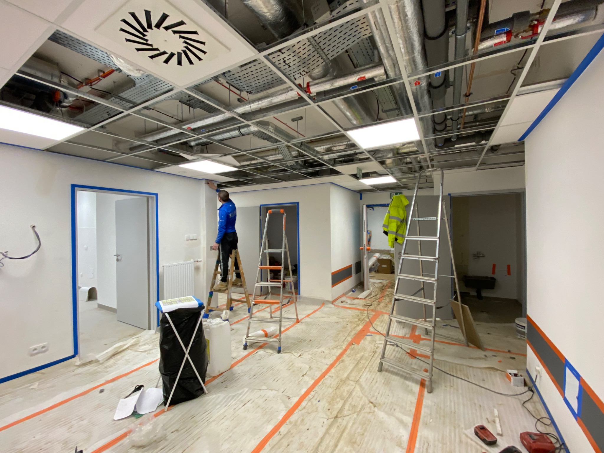 Instalacja sufitu podwieszanego z widocznymi przewodami wentylacyjnymi i rurami, pracownik na drabinie uszczelnia łączenie ściany z sufitem za pomocą białego silikonu, podłoga zabezpieczona folią...