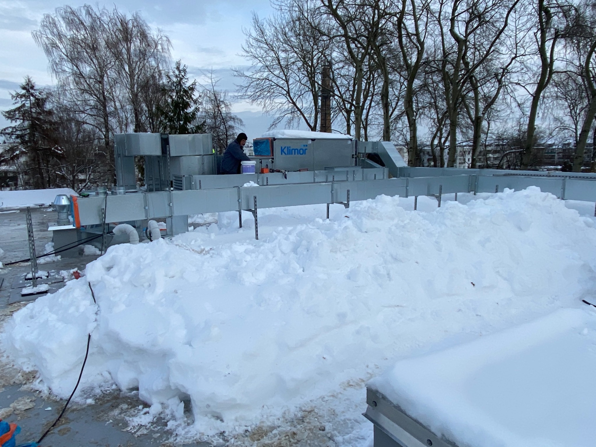 Pracownik w niebieskiej kurtce montuje system wentylacyjny Klimor na zasypanym śniegiem dachu budynku w zimowy dzień.