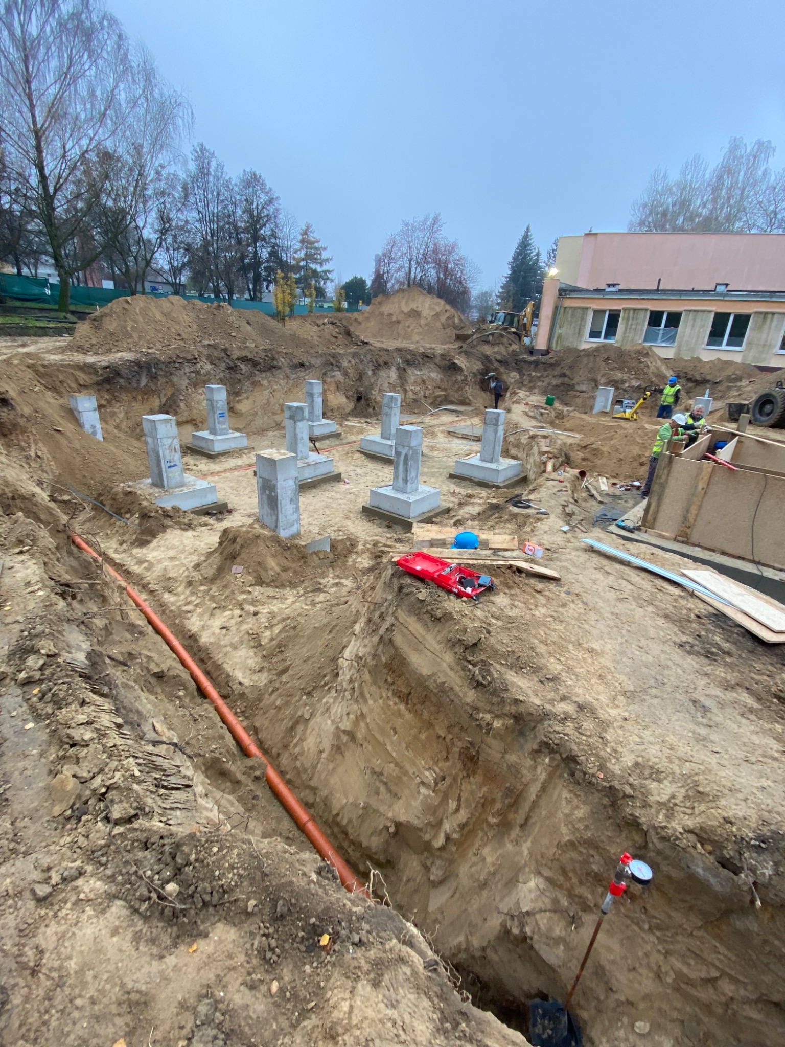 Fundamenty pod budynek w wykopie, widoczna instalacja kanalizacyjna w ziemi, pracownicy w odblaskowych kamizelkach i sprzęt budowlany w tle.