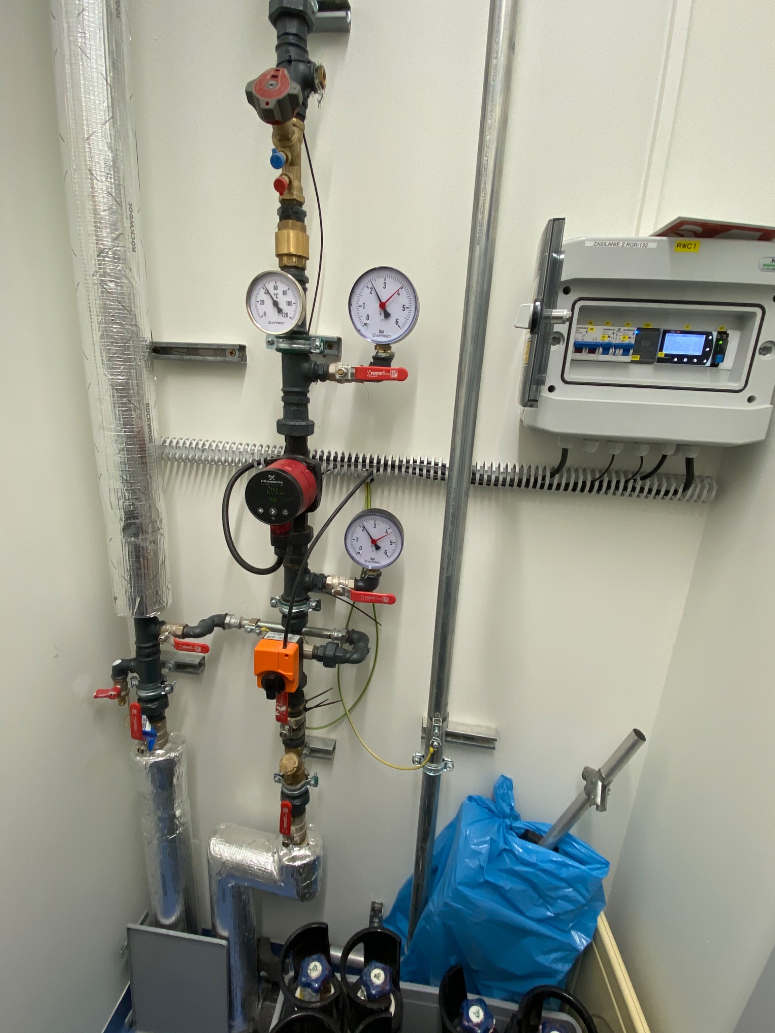 Złożona instalacja hydrauliczna z pompą obiegową Grundfos, manometrami Afriso, zaworami, rurami zaizolowanymi folią aluminiową i szafką sterowniczą ZASILANIE Z RGR/132 RWC1 na białej ścianie...