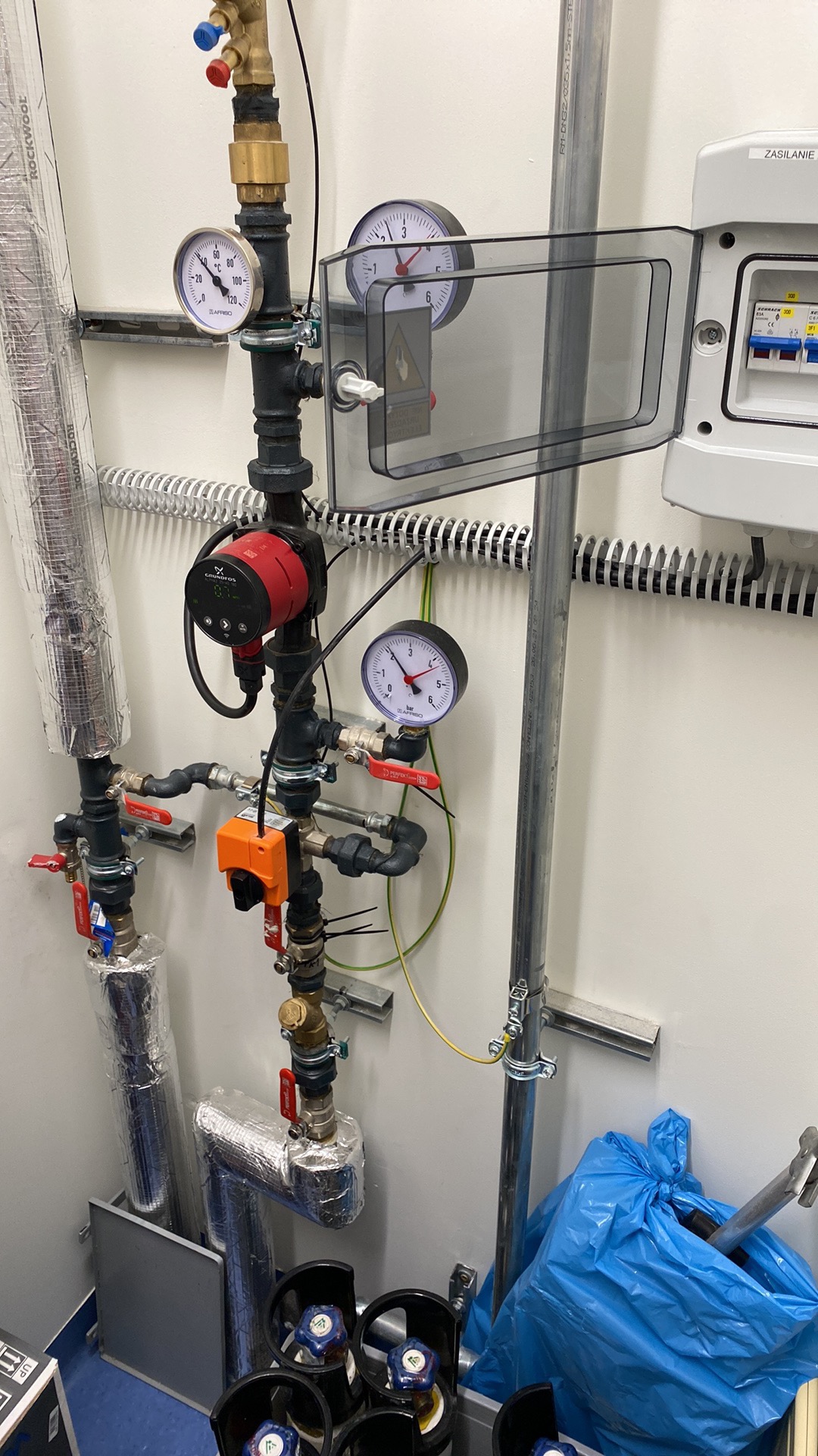 Instalacja z rur, zaworów, manometrów i pompy obiegowej w kotłowni, widoczna skrzynka elektryczna z bezpiecznikami, rury zaizolowane termicznie, niebieski worek na odpady i butle gazowe w tle.