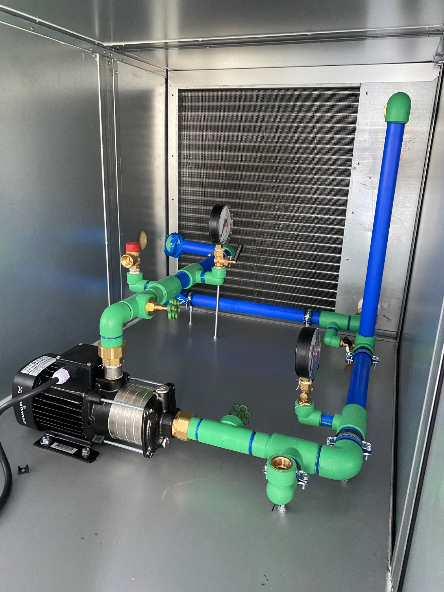 Instalacja pompy obiegowej z rurami w kolorach zielonym i niebieskim, widoczna manometry, całość umieszczona w metalowej obudowie, Zamość.