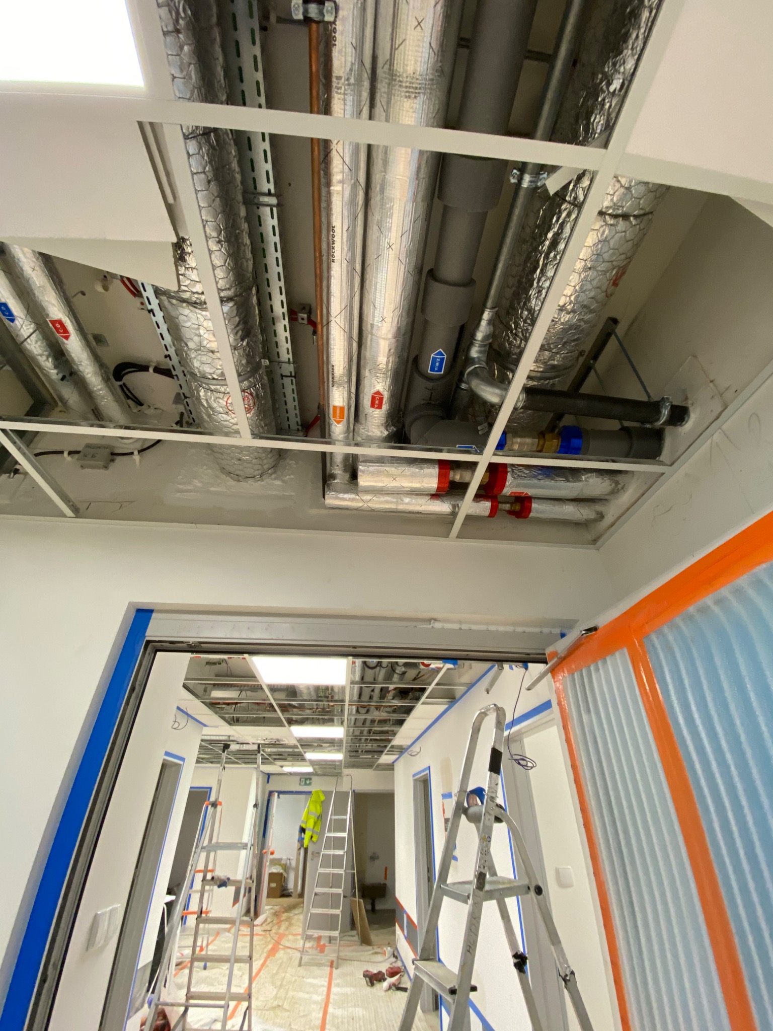 Instalacja wentylacji i rur w budynku w trakcie budowy, widoczne rury izolowane folią aluminiową, drabiny i pracownik w odblaskowej kamizelce w oddali.