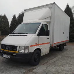 Biały samochód dostawczy Volkswagen z zabudową kontenerową, zaparkowany na betonowym placu, widoczne drzewa iglaste w tle.