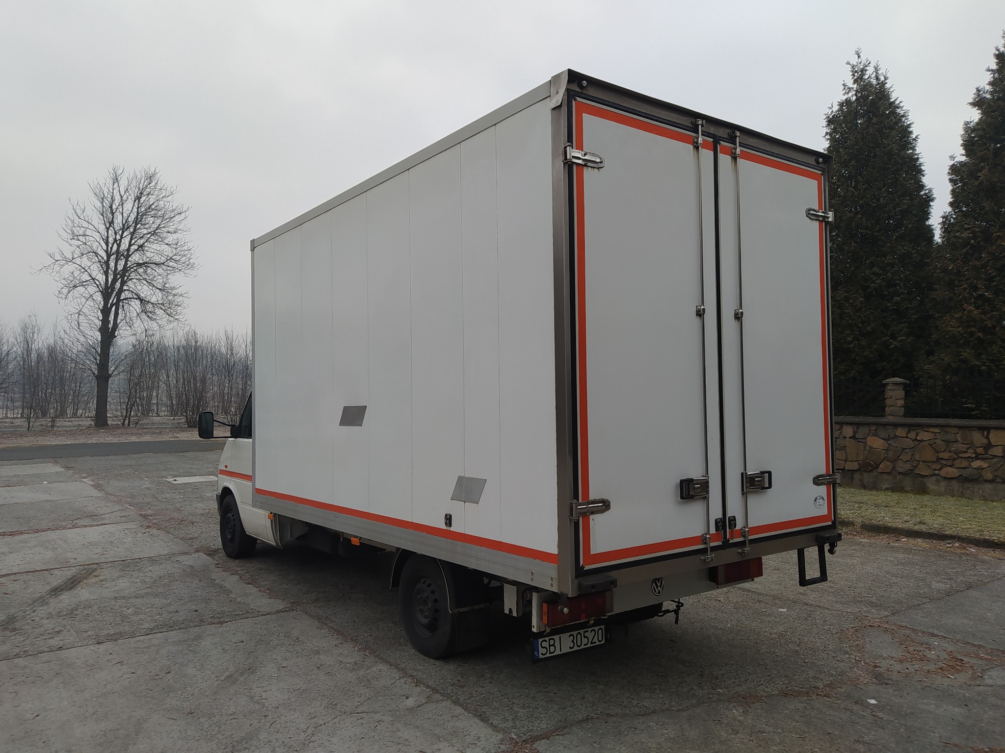 Biały samochód dostawczy z pomarańczowymi akcentami, zaparkowany na betonowym placu, z widocznym drzewem bez liści w tle i fragmentem kamiennego muru.