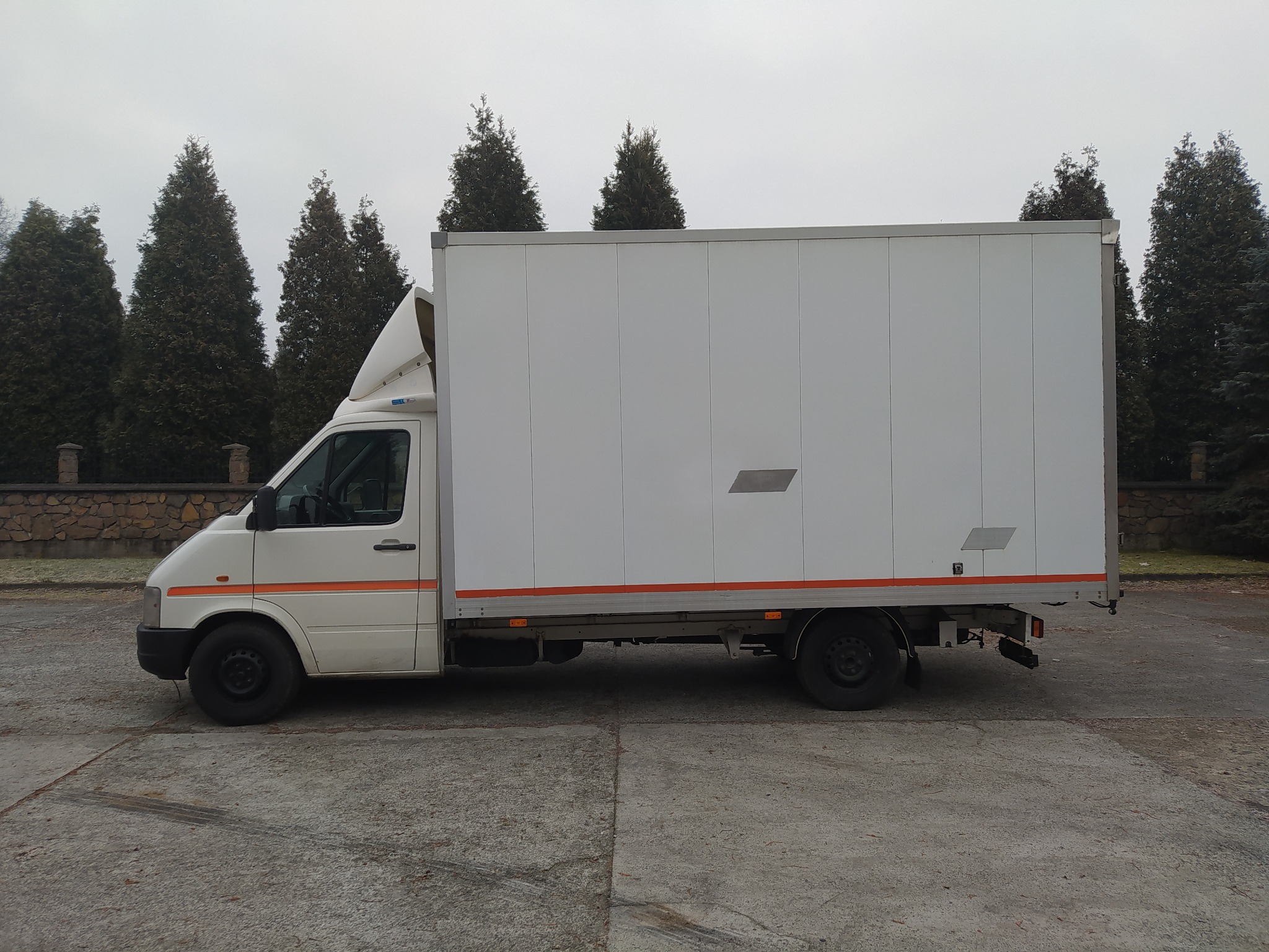 Biały samochód dostawczy z pomarańczowym paskiem na boku, zaparkowany na betonowym placu, z drzewami iglastymi w tle, sfotografowany w pochmurny dzień.
