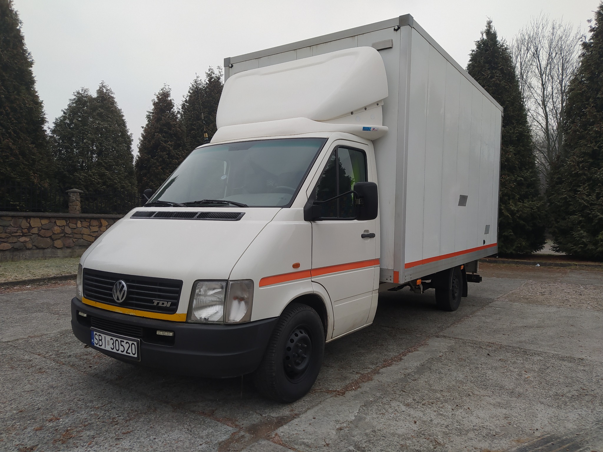 Biały samochód dostawczy Volkswagen z zabudową kontenerową, zaparkowany na betonowym placu, widoczne drzewa iglaste w tle.