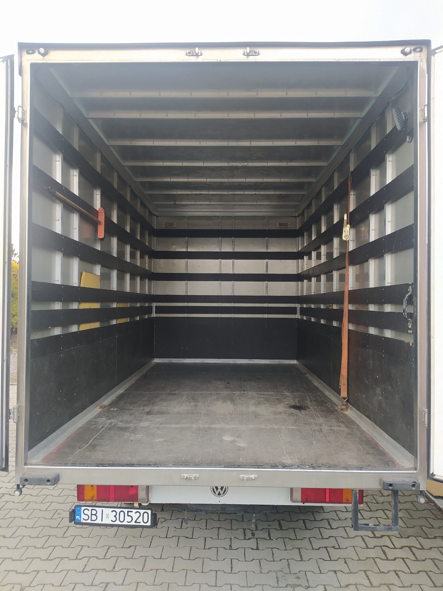 Wnętrze pustej przestrzeni ładunkowej busa z otwartymi drzwiami, widoczne zabezpieczenia ładunku na ścianach i podłodze, tablica rejestracyjna pojazdu z tyłu.