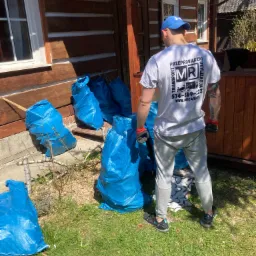 Mężczyzna w czapce i koszulce firmowej stoi tyłem do aparatu obok drewnianego domu, przed nim niebieskie worki wypełnione odpadami, prawdopodobnie po remoncie.