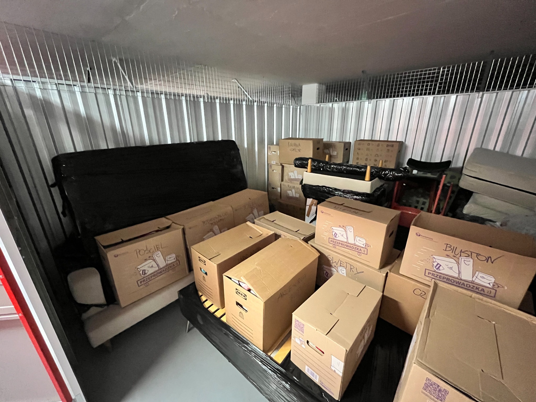Wnętrze magazynu samoobsługowego z metalowymi ścianami, wypełnione kartonami i meblami przygotowanymi do przeprowadzki, niektóre kartony opisane ręcznie z symbolami graficznymi i napisem...