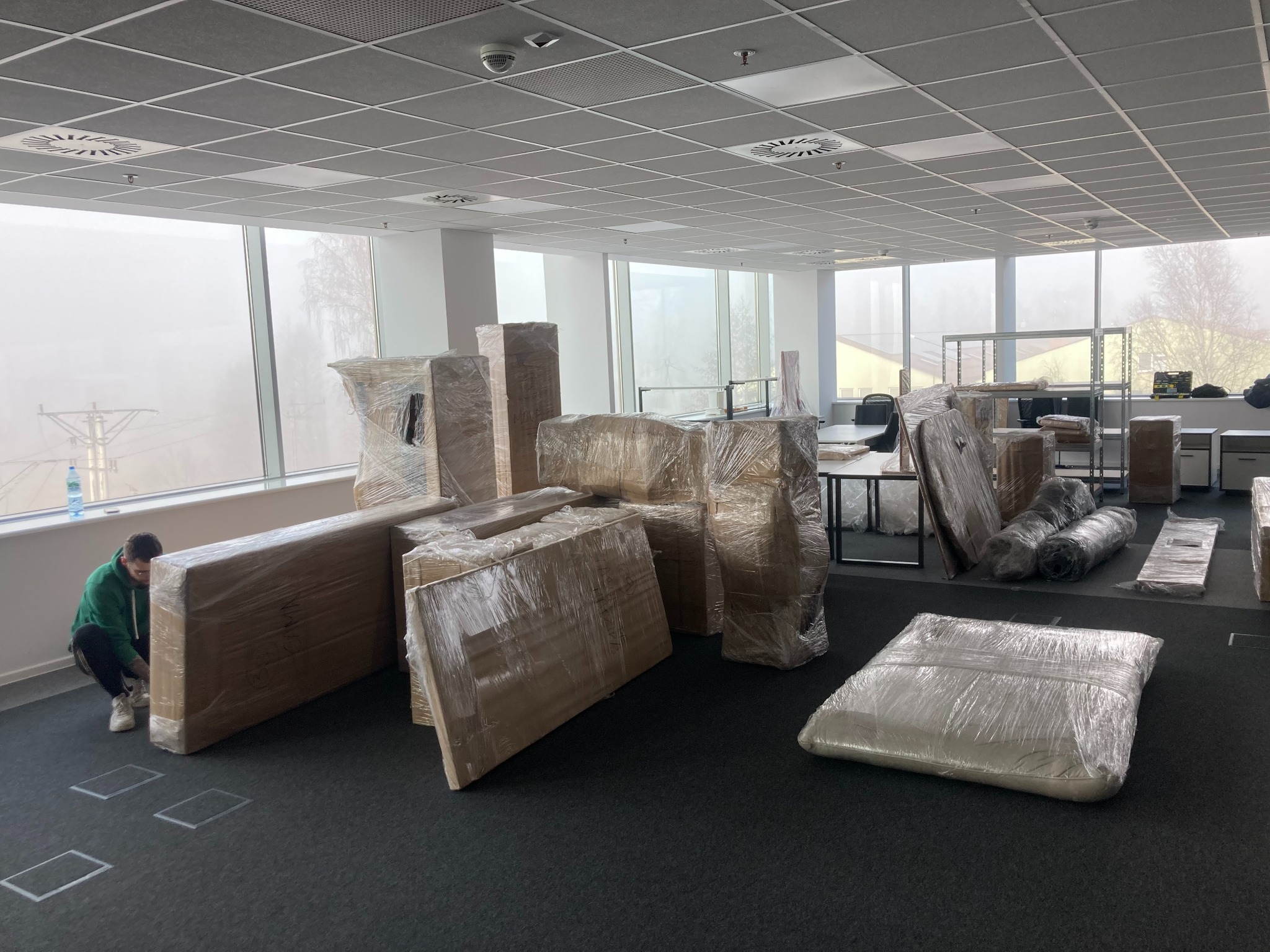 Wnętrze biura z pakunkami owiniętymi folią stretch, przygotowanymi do transportu; pracownik w zielonej bluzie kuca przy jednym z pakunków, prawdopodobnie zabezpieczając go.