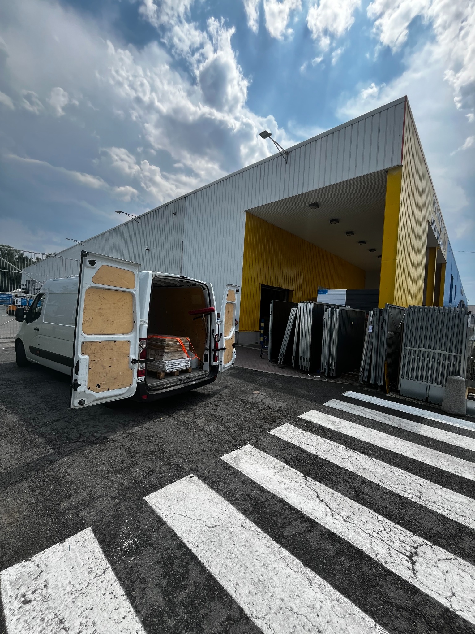 Biały van z otwartymi drzwiami bagażnika, załadowany materiałami budowlanymi zabezpieczonymi pomarańczowym pasem, zaparkowany na pasach dla pieszych przed budynkiem z żółtymi elementami.