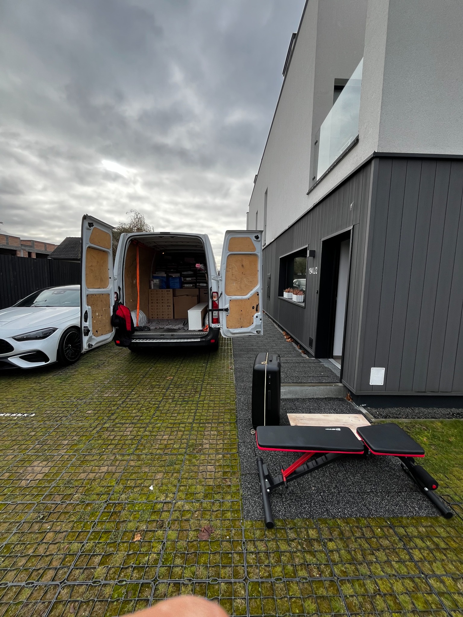 Otwarty furgon wypełniony kartonami stoi na posesji nowoczesnego domu z białymi ścianami i szarą elewacją. Obok zaparkowany biały samochód osobowy. Na pierwszym planie walizka i ławeczka do ćwiczeń.