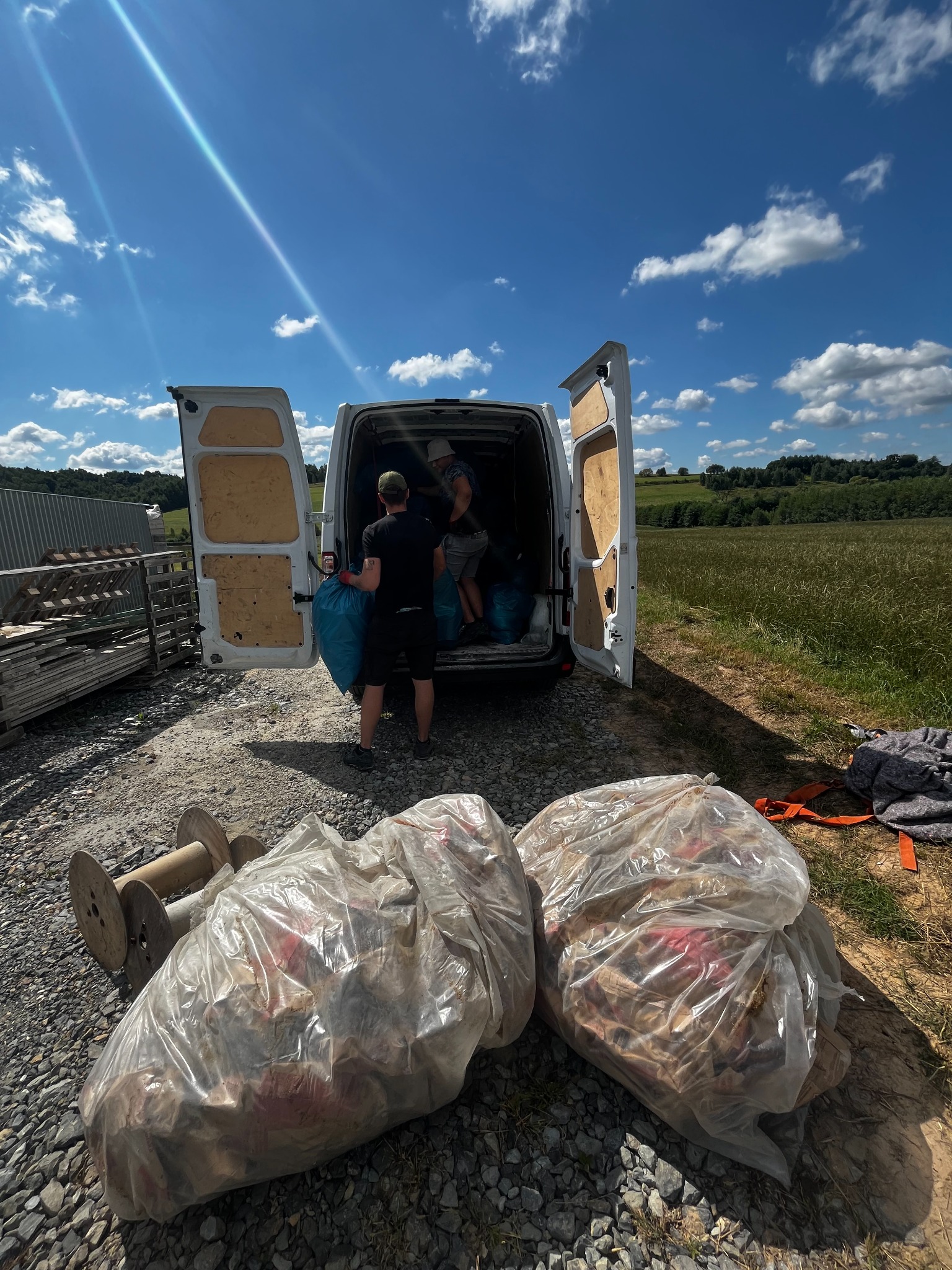 Dwóch mężczyzn ładuje worki do białego busa z otwartymi drzwiami, w tle pola i niebieskie niebo z chmurami, na pierwszym planie worki ze śmieciami i drewniane szpule na żwirowym podłożu.