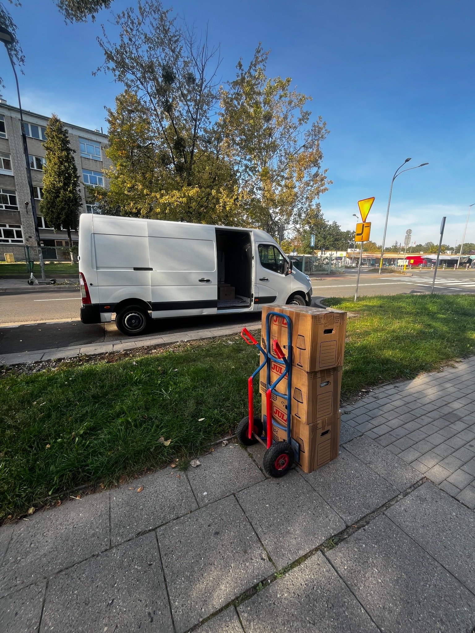 Biały van z otwartymi drzwiami, w tle budynek i drzewa. Na chodniku wózek transportowy z trzema kartonami z nadrukiem lampy.