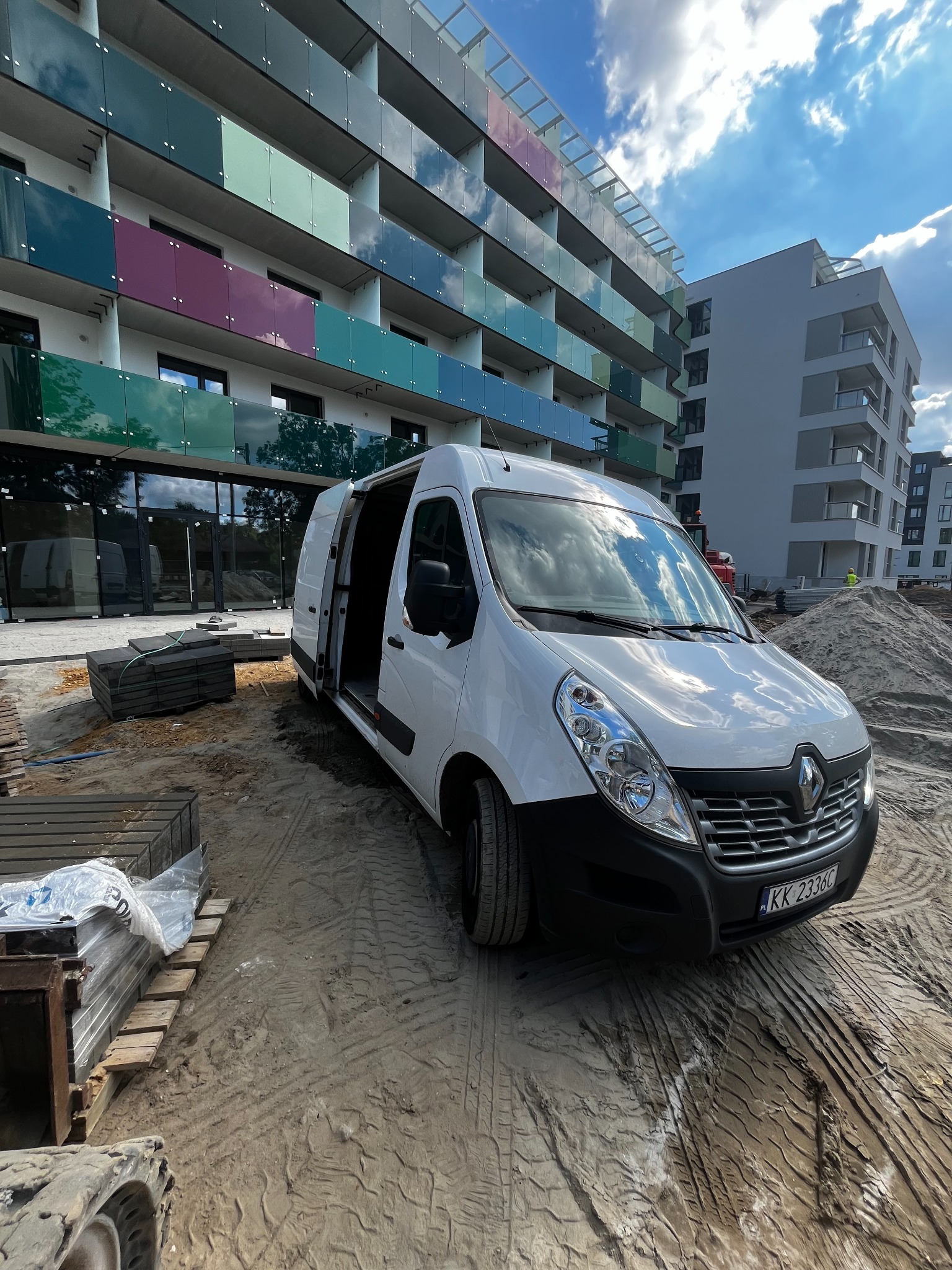 Biały Renault Master z otwartymi drzwiami na placu budowy, w tle nowoczesny budynek z kolorowymi balkonami i sterty piasku.