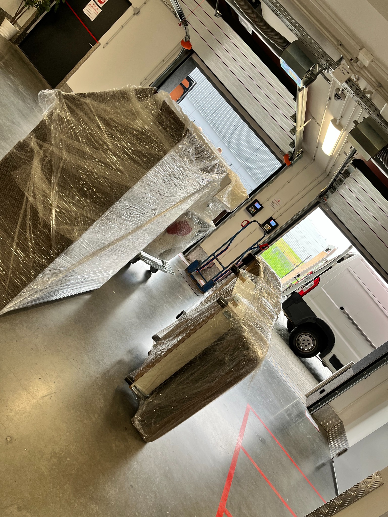 Meble owinięte folią na wózkach, przygotowane do transportu, widok z rampy załadunkowej z otwartą bramą i częściowo widocznym busem.